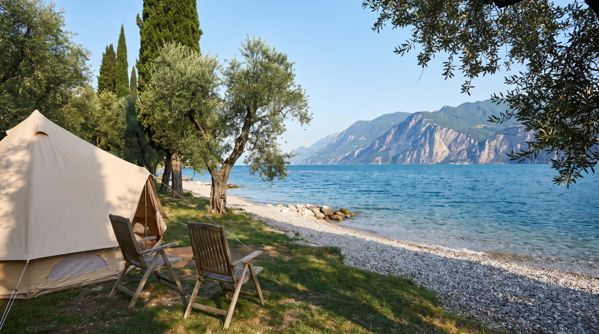 Een beige tent met houten stoelen onder bomen bij het kiezelstrand van het Gardameer, met bergen aan de overkant.